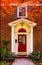 Entrance to brick house with pillars for porch and red door with autum leaves on sidewalk and colorful branches framing picture -