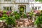 Entrance to up-scale chalet-like stucco house with brick stairs up to flower filled courtyard with fountain and trees reflected in