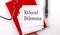 ETHICAL DILEMMA text on sticker on red notebook with pen and glasses