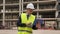 European architect in workwear and white hardhat with laptop in hands standing on building site.