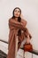 European beautiful young woman in long stylish coat in beige trendy trousers with leather brown fashionable handbag near white