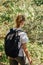 European female backpacker in white t-short walking through forest. Young hipster girl hiking with rucksack. View from back close