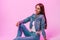 European joyful young woman fashion model with sweet smile in fashionable blue jeans clothing sits near glamorous pink wall