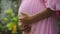 European pregnant girl in a pink dress gently touches her big stomach with her hands, stands on the back green garden