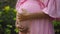 European pregnant girl in a pink dress gently touches her big stomach with her hands, stands on the back green garden