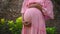 European pregnant girl in a pink dress gently touches her big stomach with her hands, stands on the back green garden