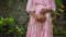 European pregnant girl in a pink dress gently touches her big stomach with her hands, stands on the back green garden