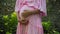 European pregnant girl in a pink dress gently touches her big stomach with her hands, stands on the back green garden