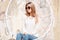 European stylish young woman hipster in sunglasses with a brilliant necklace in stylish jeans in a knitted sweater sits