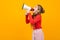 European teenager girl screaming news in a loudspeaker and stands sideways on a yellow studio background with copy space