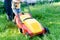 Every little help counts: image of grass trimming or lawn mover machine operating or pushing by small boy or girl and adult behind