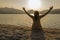 Evocative shot: woman in contemplative pose sitting on the sand with arms wide, facing the sea at sunrise or sunset on a beach.