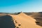 Evocative young confident woman walking on her own footprints path on the desert on top of dune with red dresson hot summer day