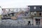 An excavator demolishes a residential building near a roadway. The floors are broken, litter is lying around, dust is flying.