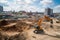 excavator digging into pile of dirt, with view of the bustling construction site in the background