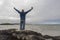 Excited bald man standing on a rock by the ocean, hands up in the air, beautiful and calm scenery in the background. West coast of