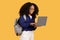 Excited black lady student in glasses with backpack, using laptop and smiling, against yellow background