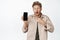 Excited blond curly guy pointing finger at mobile phone screen, looking surprised, showing awesome news online, standing