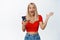Excited blond girl reacts to mobile phone notification, holding smartphone and shouting with joy, standing over white