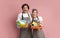 Excited Couple Holding Baskets With Cleaning Supplies, Posing Over Pink Background