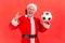 Excited crazy elderly man with gray beard wearing santa claus costume standing with soccer ball in hands and showing rock sign to