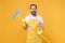 Excited crazy young man househusband in apron rubber gloves hold in hands broom while doing housework isolated on yellow