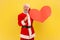 Excited elderly man with gray beard wearing santa claus costume keeps mouth open, showing big red heart and v sign, celebrating