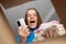 Excited extremely happy joyful cheerful dark haired woman in blue shirt looking inside cardboard box, holding smart phone and