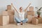 Excited extremely happy woman wearing white T-shirt sitting on the floor near cardboard boxes with stuff, celebrating relocation