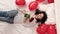 Excited Girl falling on bed red heart shape balloons with bouquet of flowers. Woman rejoices gift on valentines day