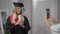 Excited graduate daughter posing with rolled diploma as mother taking photos on smartphone indoors. Portrait of happy