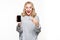 Excited happy blond woman in sweater showing blank smartphone screen and pointing on it while looking at camera with open mouth.