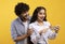 Excited indian couple looking at smartphone screen, using new app or reading cool news, standing over yellow background