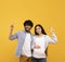 Excited indian pregnant lady and her husband pointing up at free space, showing amazing sale or offer, yellow background