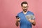 Excited man in casual t-shirt holding lots of money in dollar currencys and cell phone in hands isolated over pink wall