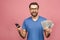 Excited man in casual t-shirt holding lots of money in dollar currencys and cell phone in hands isolated over pink wall