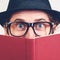 Excited nerd hiding behind books. Playful happy young man in funny glasses and vintage hat holding a book. Nerd hipster guy in hat