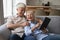 Excited older grandparents hug on couch laugh using digital touchpad