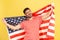 Excited patriotic man with beard in striped t-shirt standing holding in hands flag of united states of america and screaming,