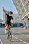 Excited professional female cyclist in black cycling garment and protective gear smiling, raising her arm, looking