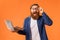 Excited redhaired bearded programmer guy holds laptop over orange background