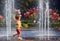 Excited redhead baby boy having fun between water jets, in fountain. Summer in the city