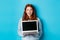 Excited redhead female freelancer showing laptop screen, staring at camera amazed, standing with computer against blue