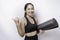Excited sporty Asian woman wearing sportswear pointing at the copy space beside her while carrying yoga mat, isolated by white