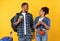 Excited Travelers Spouses Using Mobile Banking Standing Over Yellow Background