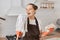 Excited woman with dark hair wearing white t shirt and brown apron, holding mop and cloth in hands, housekeeper singing while