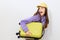 an excited woman is standing sideways on a white background in a purple tracksuit and a yellow cap, clutching a suitcase