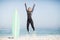 Excited woman in wetsuit jumping next to her surfboard