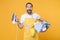 Excited young man househusband in apron hold basket with clean clothes, iron doing housework isolated on yellow