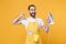 Excited young man househusband in apron hold in hands iron while doing housework isolated on yellow background studio